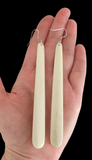 Long, thin, off-white earrings held in a hand against a black background