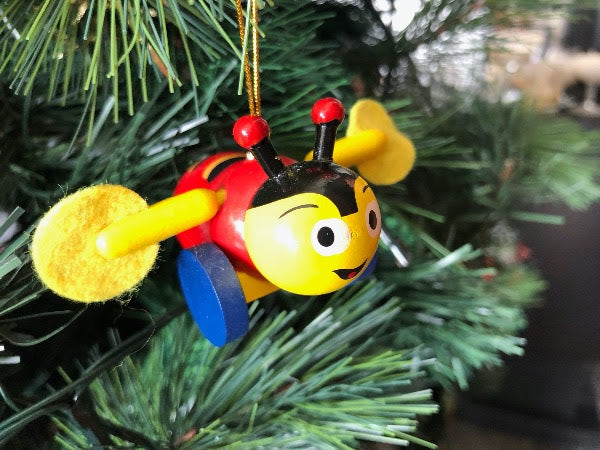 A small hanging decoration resembling a buzzy bee with yellow and red coloring and a blue patterned abdomen, attached to a Christmas tree.