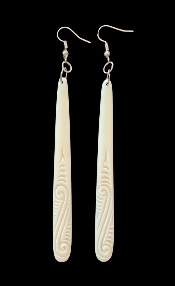 A pair of long, narrow white carved dangle earrings with silver fishhook wires, shown against a solid black background.