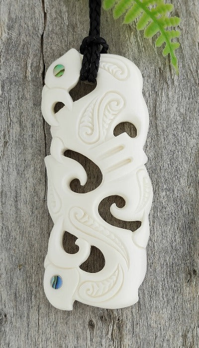 A carved bone necklace with floral and geometric patterns and Paua eyes, displayed on a wooden surface, with an adjustable black cord.