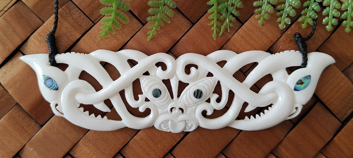 A Maori bone carving pendant featuring a Manaia and Tiki design with eyes made from Paua Shell, displayed on a black waxed cord.
