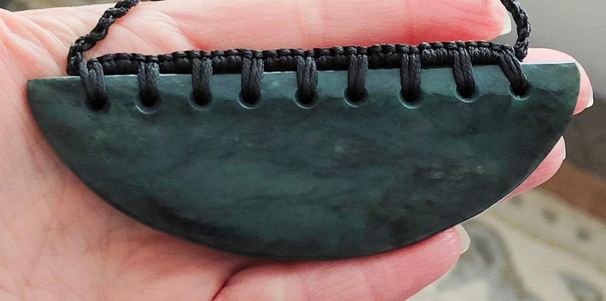 A greenstone breast plate pendant with a black adjustable slipknot cord, held between fingers.
