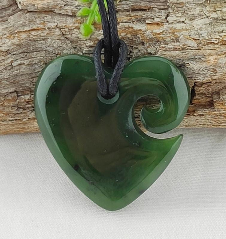 A greenstone heart-shaped pendant with a Koru design, attached to a black slipknot cord, displayed against a wooden background. Main image