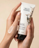 Hand holding a tube of Wild Ferns hand cream with a visible smear of cream on the other hand against a beige background.