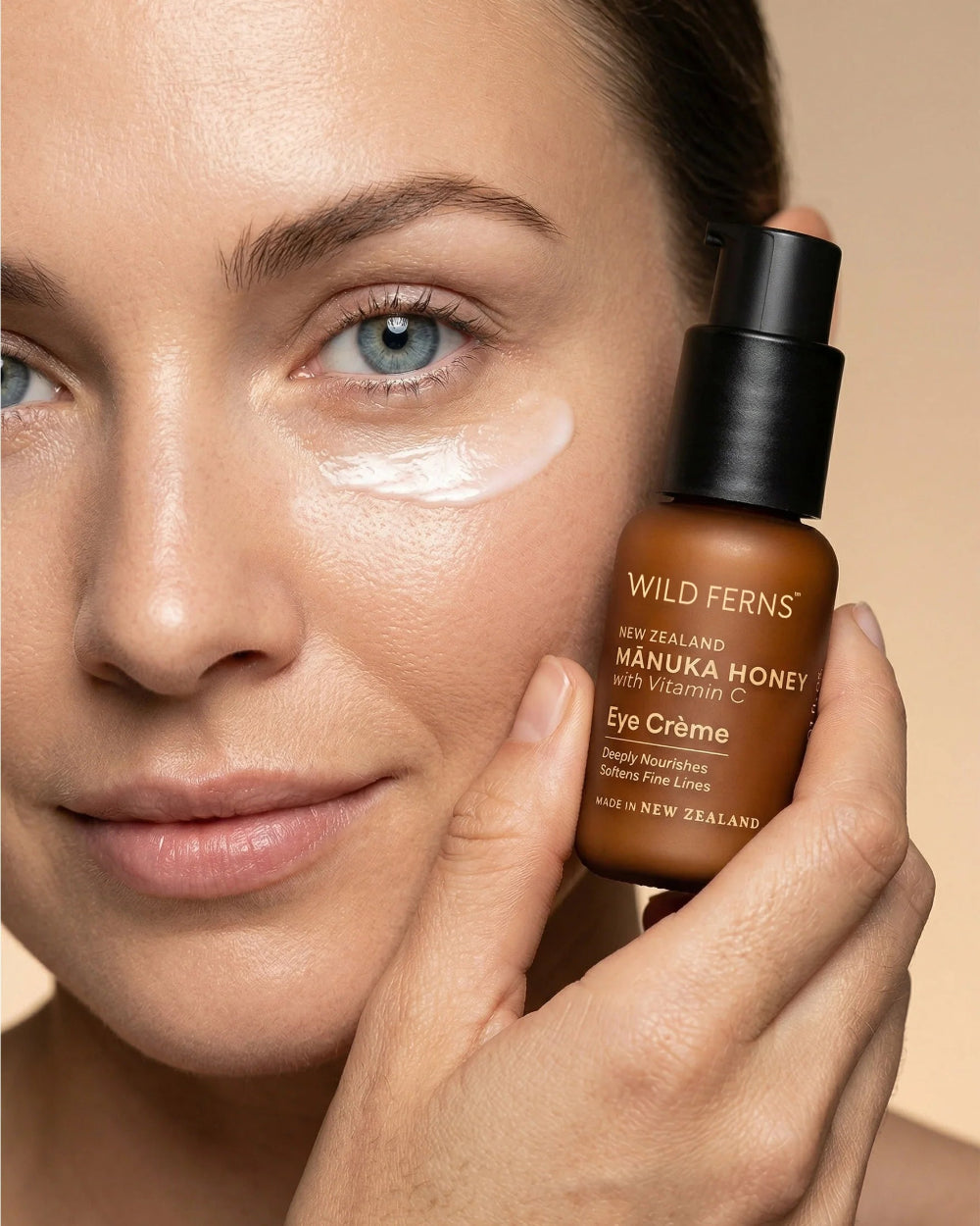 Woman holding a bottle of Wild Ferns Manuka Honey Eye Crème near her face.