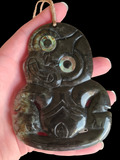 A carved dark green stone pendant with mother-of-pearl iridescent eye details, held in a person's hand against a solid black background.