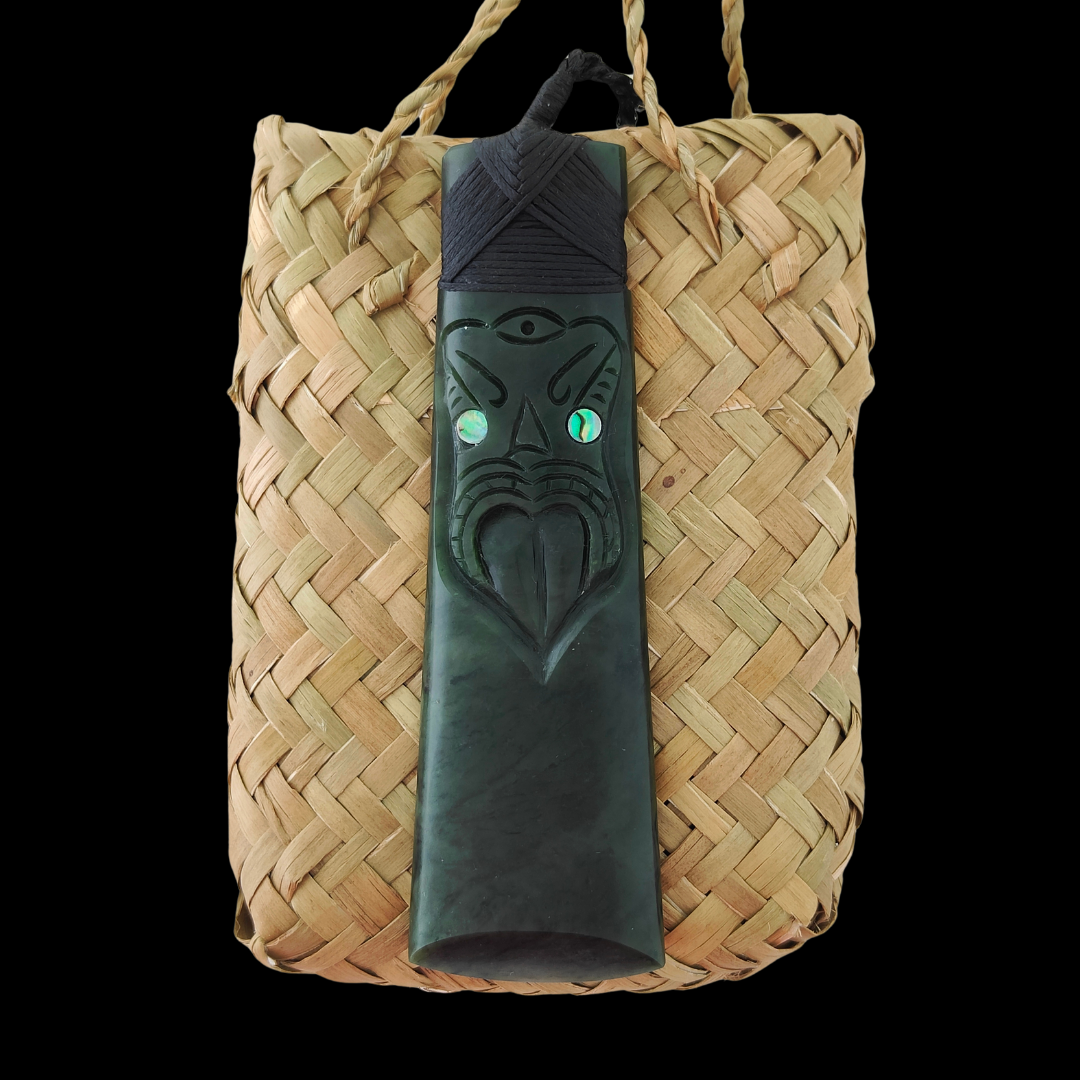 A greenstone carving of a Toki and Mask with paua eyes displayed on a woven kete bag.