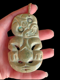 A carved stone pendant with a tribal face design, held in a hand against a solid black background. The pendant is made of a polished light green stone with iridescent shell inlays and a cord loop at the top.
