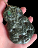 A polished dark green carved stone pendant with two iridescent green-blue eye motifs, held in a person's hand against a solid black background.