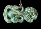 A carved green stone pendant with iridescent shell-like eye details and a braided cord loop at the top, shown against a solid black background.