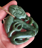 A green jadeite carved pendant with a stylized face and flowing organic lines, held in a person's hand against a black background.