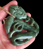 A green jadeite carved pendant with a stylized face and flowing organic lines, held in a person's hand against a black background.