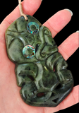 A carved green jade pendant with abalone shell accents, held in a hand against a solid black background.