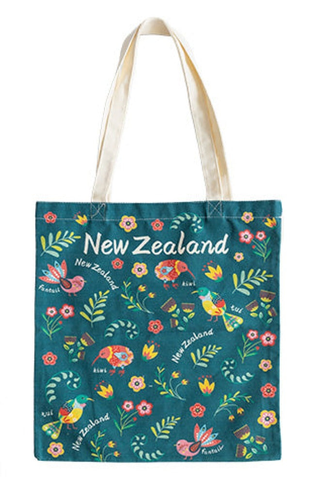 A tote bag with a dark teal background featuring a colorful New Zealand-themed pattern of birds, flowers, leaves, and the text 'New Zealand' in white. The bag has off-white canvas shoulder straps and is photographed against a plain white background.