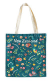 A tote bag with a dark teal background featuring a colorful New Zealand-themed pattern of birds, flowers, leaves, and the text 'New Zealand' in white. The bag has off-white canvas shoulder straps and is photographed against a plain white background.