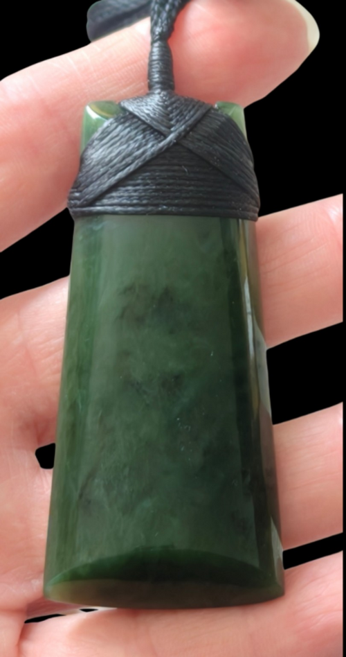 A polished dark green stone pendant held in a hand, featuring a wrapped black cord top 