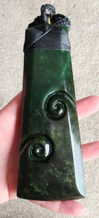 A dark green carved stone pendant with spiral motifs and a black cord wrap, held in a person's hand against a neutral background.