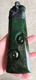 A dark green carved stone pendant with spiral motifs and a black cord wrap, held in a person's hand against a neutral background.