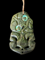 A carved green stone pendant or wall hanging with a stylized face and two iridescent circular inlays, suspended by a tan cord against a black background.