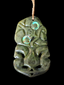 A carved green stone pendant or wall hanging with a stylized face and two iridescent circular inlays, suspended by a tan cord against a black background.