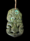 A carved green stone pendant or wall hanging with a stylized face and two iridescent circular inlays, suspended by a tan cord against a black background.