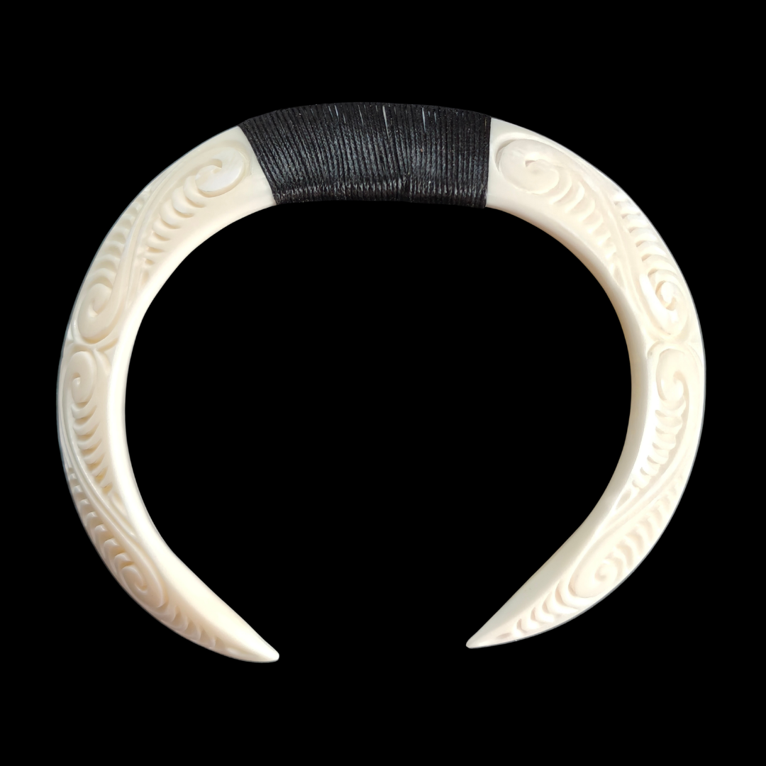 An off-white carved pig tusk necklace with intricate scrollwork patterns and a black braided cord wrapped around the top center, shown against a solid black background.
