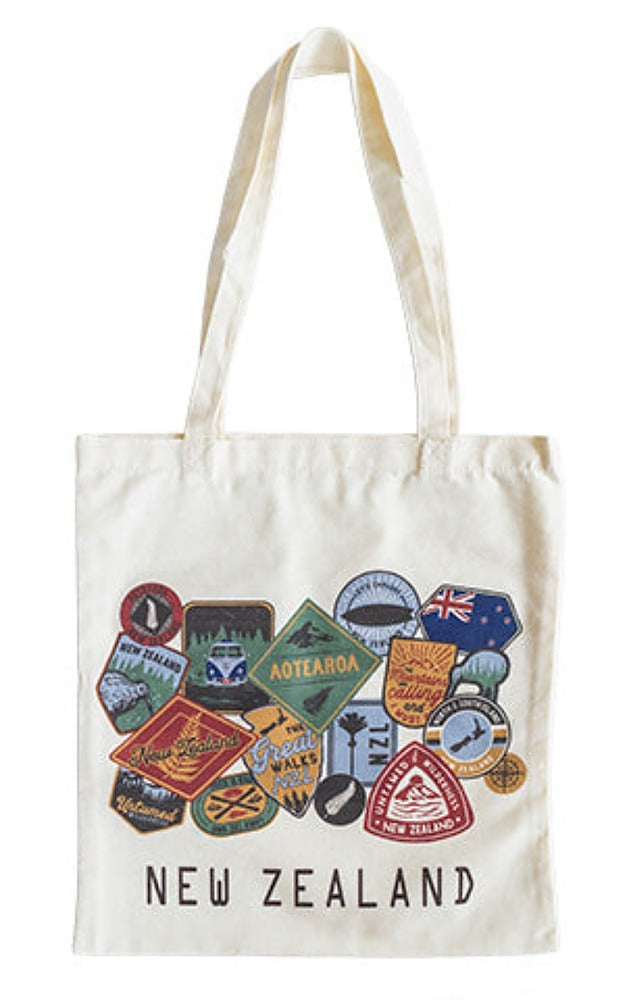 An off-white canvas tote bag with long handles, featuring a collage of colorful New Zealand travel patches and the text 'NEW ZEALAND' printed in black at the bottom.