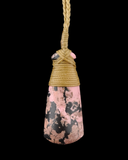 A conical pink stone pendant with black and brown marbled patterns, wrapped at the top with tan cord and suspended by a matching tan rope against a solid black background.