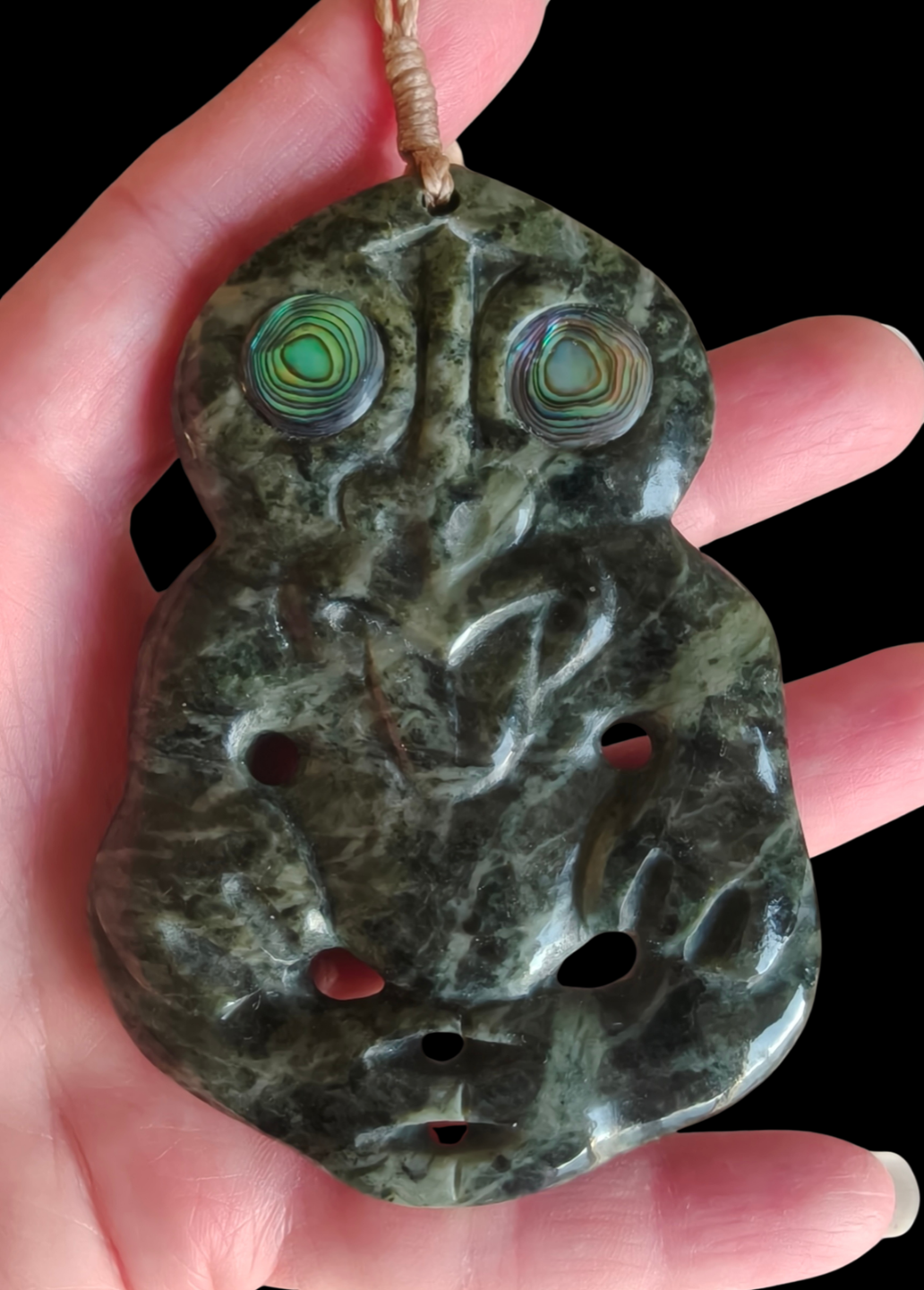 A carved green stone pendant with abalone shell eyes, held in a hand against a black background. Main image