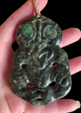 A carved green stone pendant with abalone shell eyes, held in a hand against a black background.