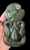 A carved green stone or wood pendant with a black cord, held in a hand against a black background. The pendant features a stylized face with large iridescent circular eye inlays.