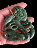 A green jade-like carved pendant with a loop at the top, held in a hand against a black background. The pendant is shaped like a stylized human figure and has a small tan cord loop.