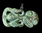 A carved green stone pendant with a dark cord loop at the top, featuring abstract facial carvings and iridescent inlay around the eye areas, shown against a solid black background.