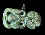 A carved green stone pendant with a dark cord loop at the top, featuring abstract facial carvings and iridescent inlay around the eye areas, shown against a solid black background.
