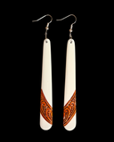 A pair of long, tapered white dangle earrings with silver fishhook wires and a carved brown geometric pattern near the bottom, shown against a solid black background.