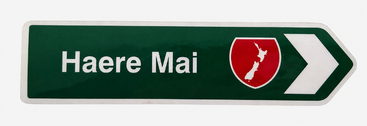 A green directional road sign pointing right with the white text 'Haere Mai' and a red shield emblem containing the white outline of New Zealand, on a light gray background.