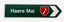 A green directional road sign pointing right with the white text 'Haere Mai' and a red shield emblem containing the white outline of New Zealand, on a light gray background.