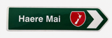 A green directional road sign pointing right with the white text 'Haere Mai' and a red shield emblem containing the white outline of New Zealand, on a light gray background.
