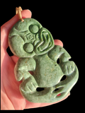 A hand holds a carved green jade pendant with a stylized figure design and a gold-colored loop at the top, set against a solid black background.