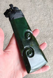 A hand holds a polished dark green jade pendant with carved spiral motifs and a black cord tie, against a neutral concrete background.