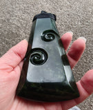 A hand holding a polished dark green carved stone object with spiral motifs, photographed outdoors against a textured gray ground.