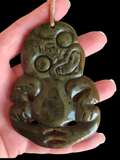 A hand holds a carved jade pendant against a solid black background. The pendant is olive green with natural speckles and a hanging cord.