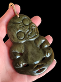 A hand holding a carved jadeite pendant shaped like a seated figure, shown against a solid black background.