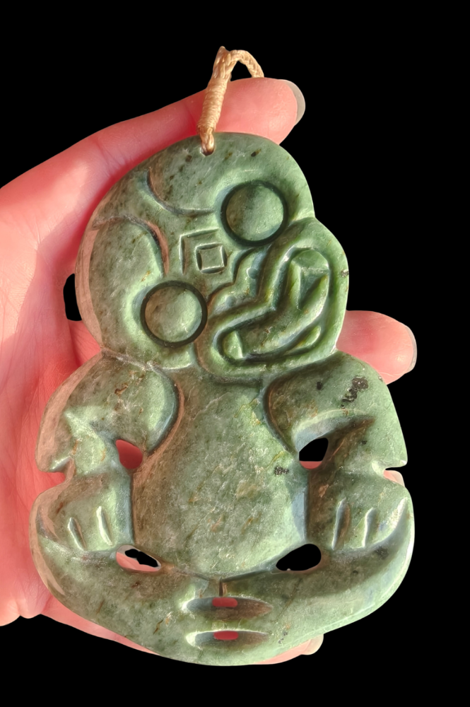 A hand holding a carved green jade pendant with a stylized face and traditional tribal motifs, set against a solid black background.