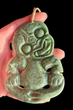 A hand holding a carved green jade pendant with a stylized face and traditional tribal motifs, set against a solid black background.