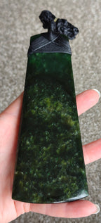 A hand holding a polished dark green stone pendant or tiki toa with a black cord tie, set against a neutral gray carpeted background.