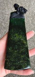 A hand holding a polished dark green stone pendant or tiki toa with a black cord tie, set against a neutral gray carpeted background.
