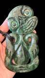 A green carved stone or jade pendant with intricate tribal-style engravings and iridescent eye inlays, held in a person's hand against a black background.