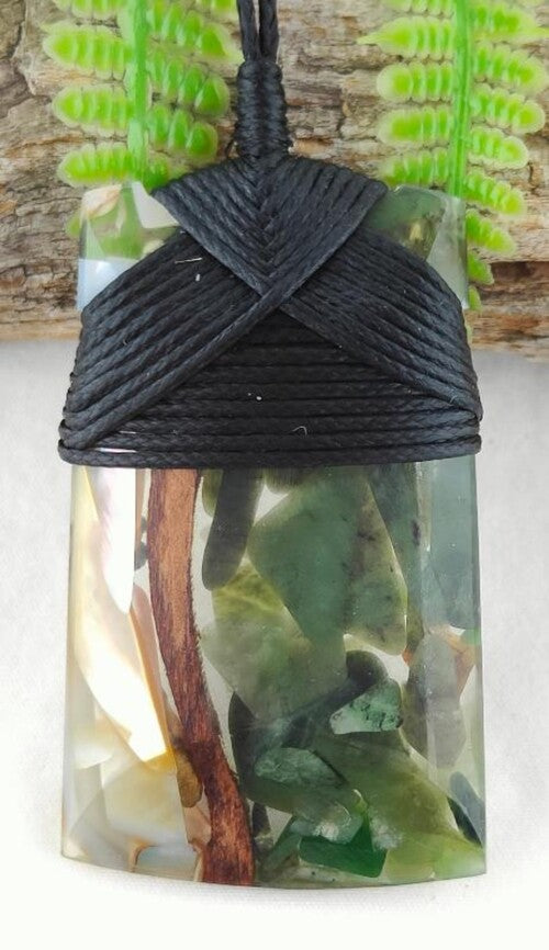 A resin pendant featuring a black braided wrap closure and a clear resin body filled with natural elements including green leaves, brown twigs, and white flower petals, shown against a background of faux ferns and wood. Main image