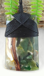 A resin pendant featuring a black braided wrap closure and a clear resin body filled with natural elements including green leaves, brown twigs, and white flower petals, shown against a background of faux ferns and wood.